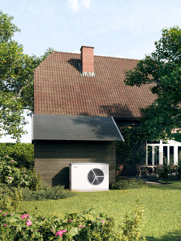 Weiße Wärmepumpe steht neben einem kleinen Gartenhaus vor einem Wohnhaus mit rotem Dach.